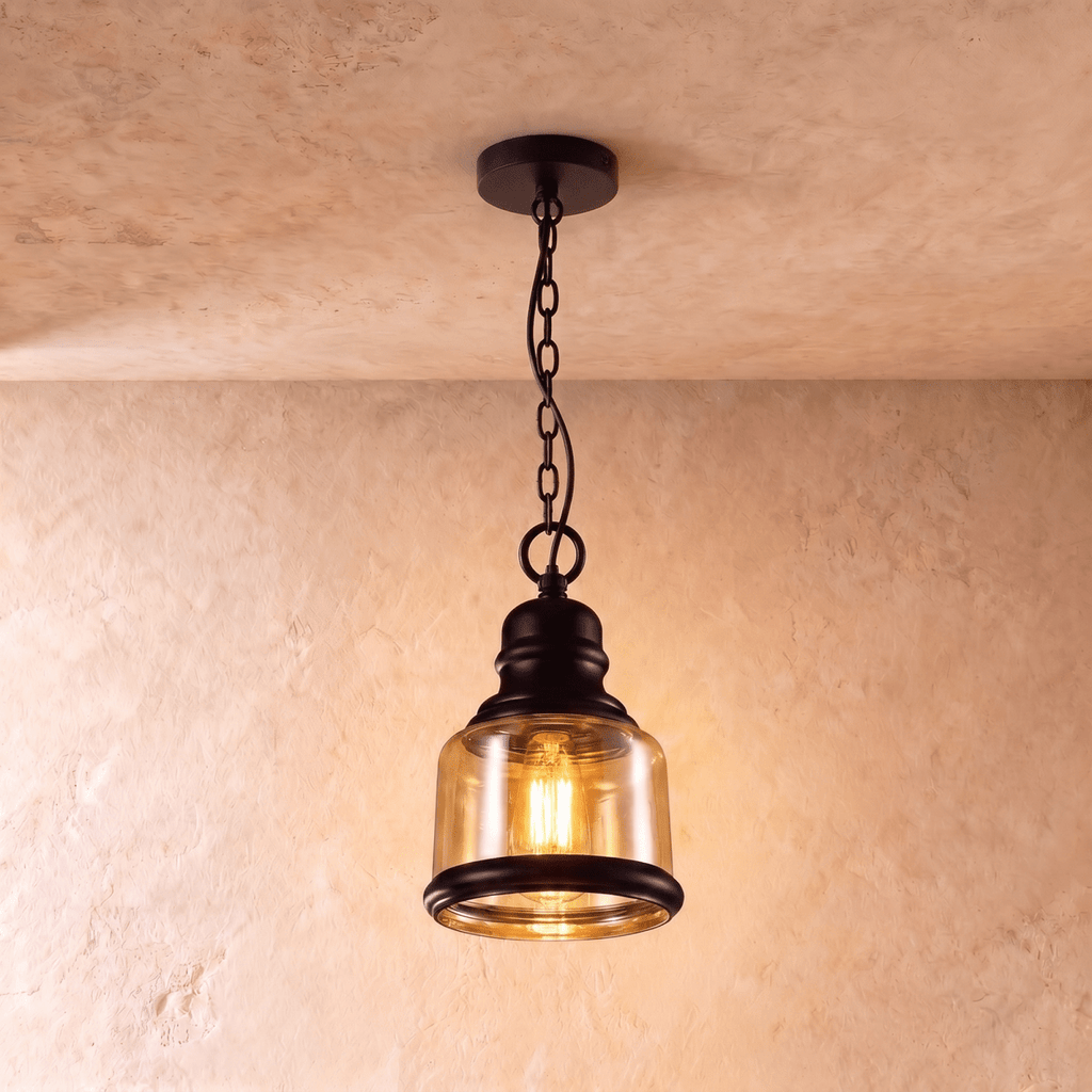 Clérachic pendant with a short amber glass cylinder shade, black iron collar and chain, glowing warm against plaster.