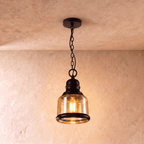 Clérachic pendant with a short amber glass cylinder shade, black iron collar and chain, glowing warm against plaster.