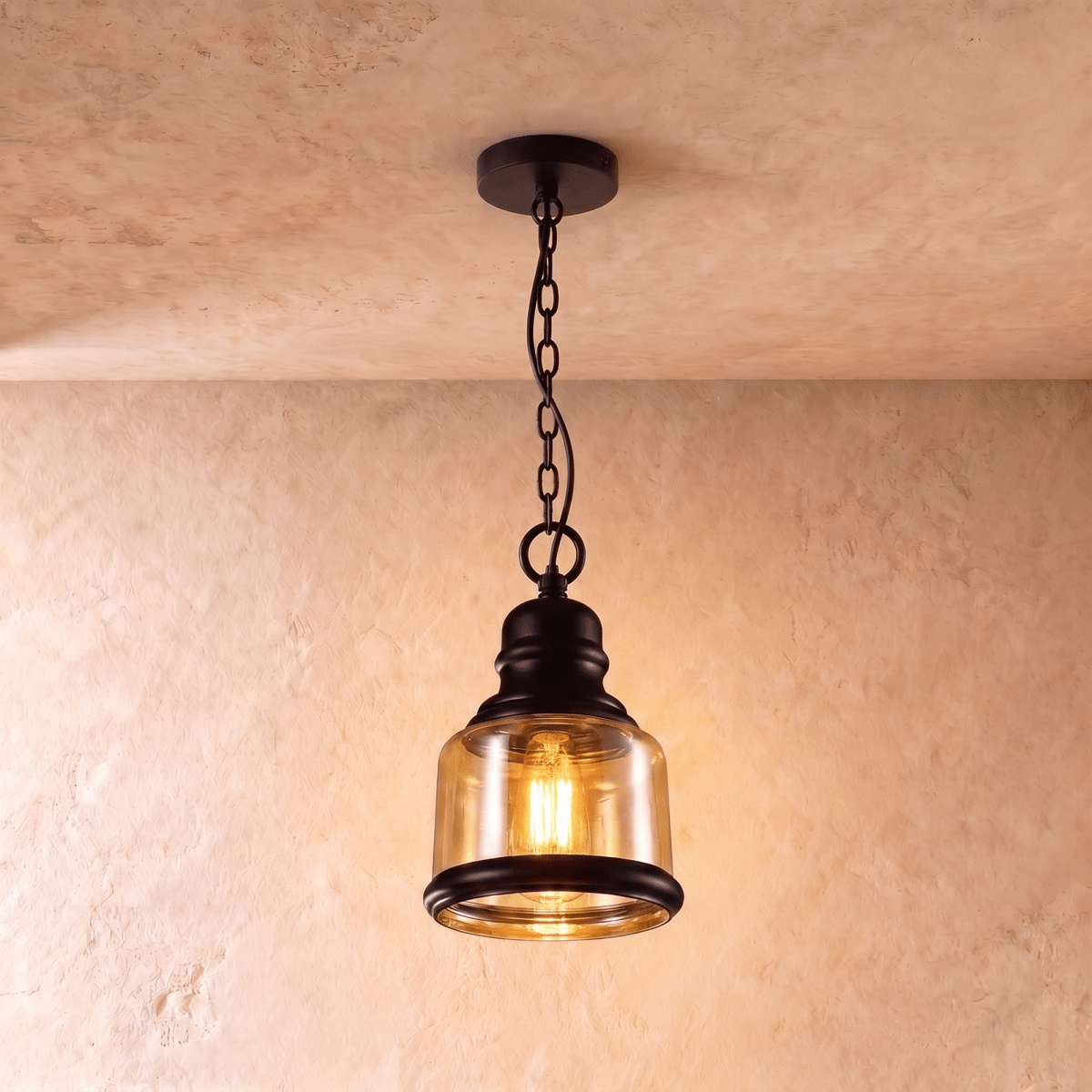 Clérachic pendant with a short amber glass cylinder shade, black iron collar and chain, glowing warm against plaster.