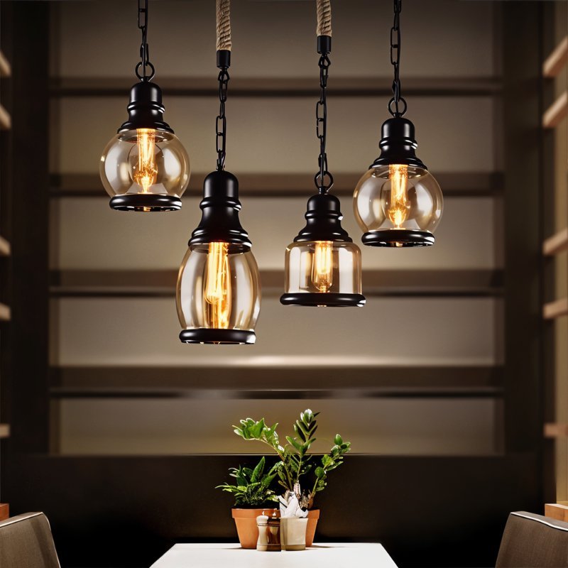 Four Clérachic amber glass pendants on black chain and rope in several shapes, glowing warm over a dining table.