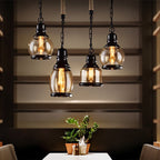 Four Clérachic amber glass pendants on black chain and rope in several shapes, glowing warm over a dining table.