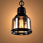 Clérachic amber glass cylinder pendant with a black iron cap and rim, its filament bulb lit warm on a golden backdrop.