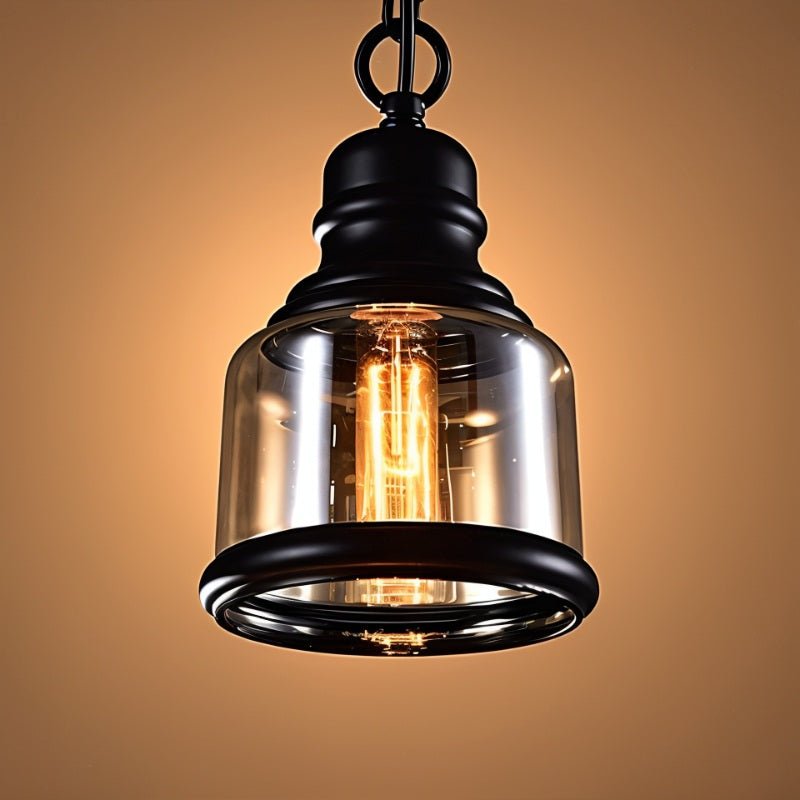 Clérachic amber glass cylinder pendant with a black iron cap and rim, its filament bulb lit warm on a golden backdrop.