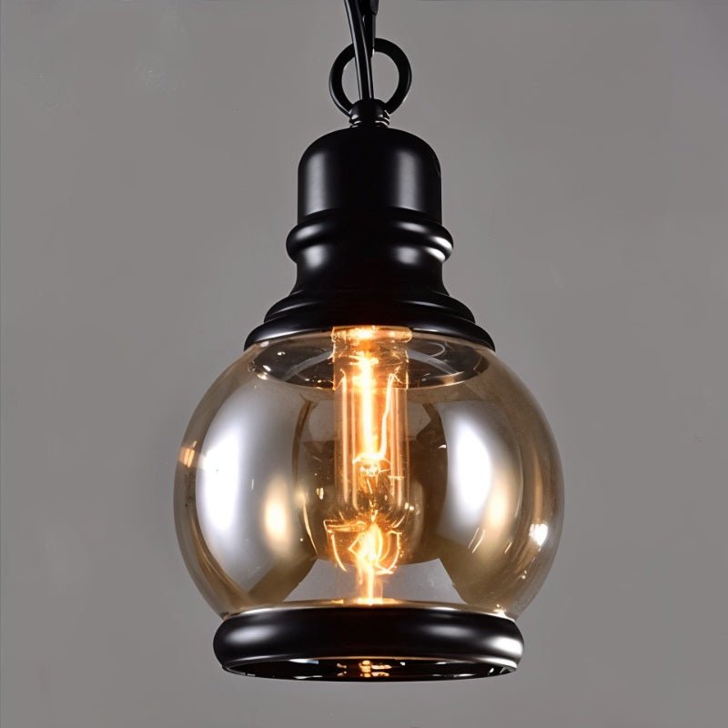 Clérachic round amber glass globe pendant with black cap and base ring, a filament bulb glowing inside, on grey.
