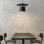 Clérachic black saucer-and-drum pendant over a dark table, warm light falling between two chairs.