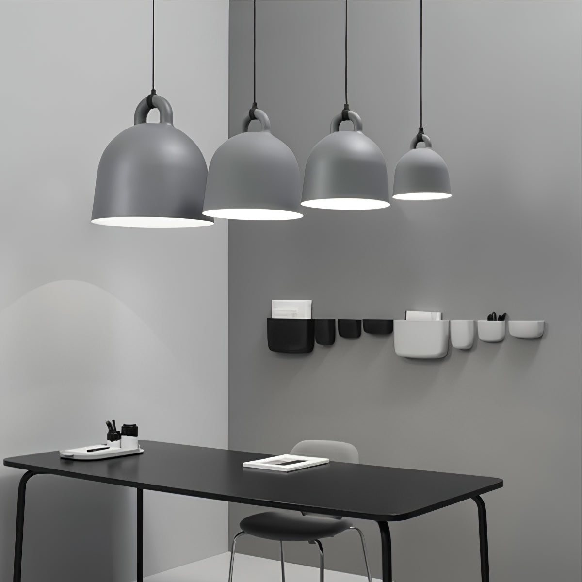 Four grey Clérachic bell pendants in graduated sizes hang over a black desk, with small wall pockets on the grey wall behind.