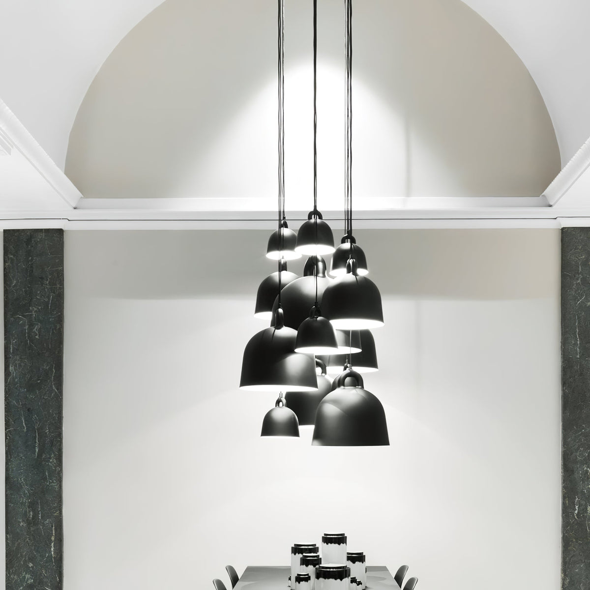 A cluster of black Clérachic bell pendants hangs at different heights in a tall, white arched room with dark green stone panels.