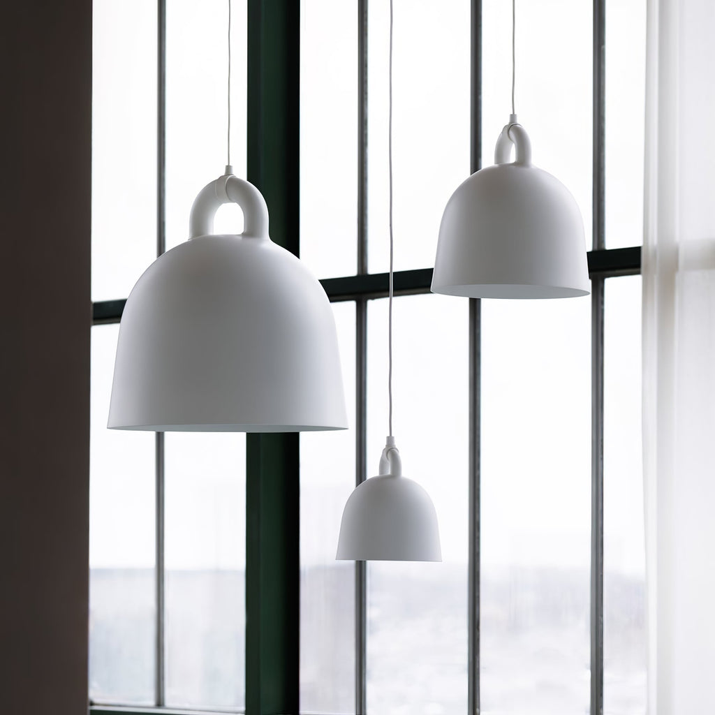 Large and small white Clérachic bell pendants hang by tall windows, with a sheer curtain and a green frame beside them.
