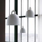 Large and small white Clérachic bell pendants hang by tall windows, with a sheer curtain and a green frame beside them.