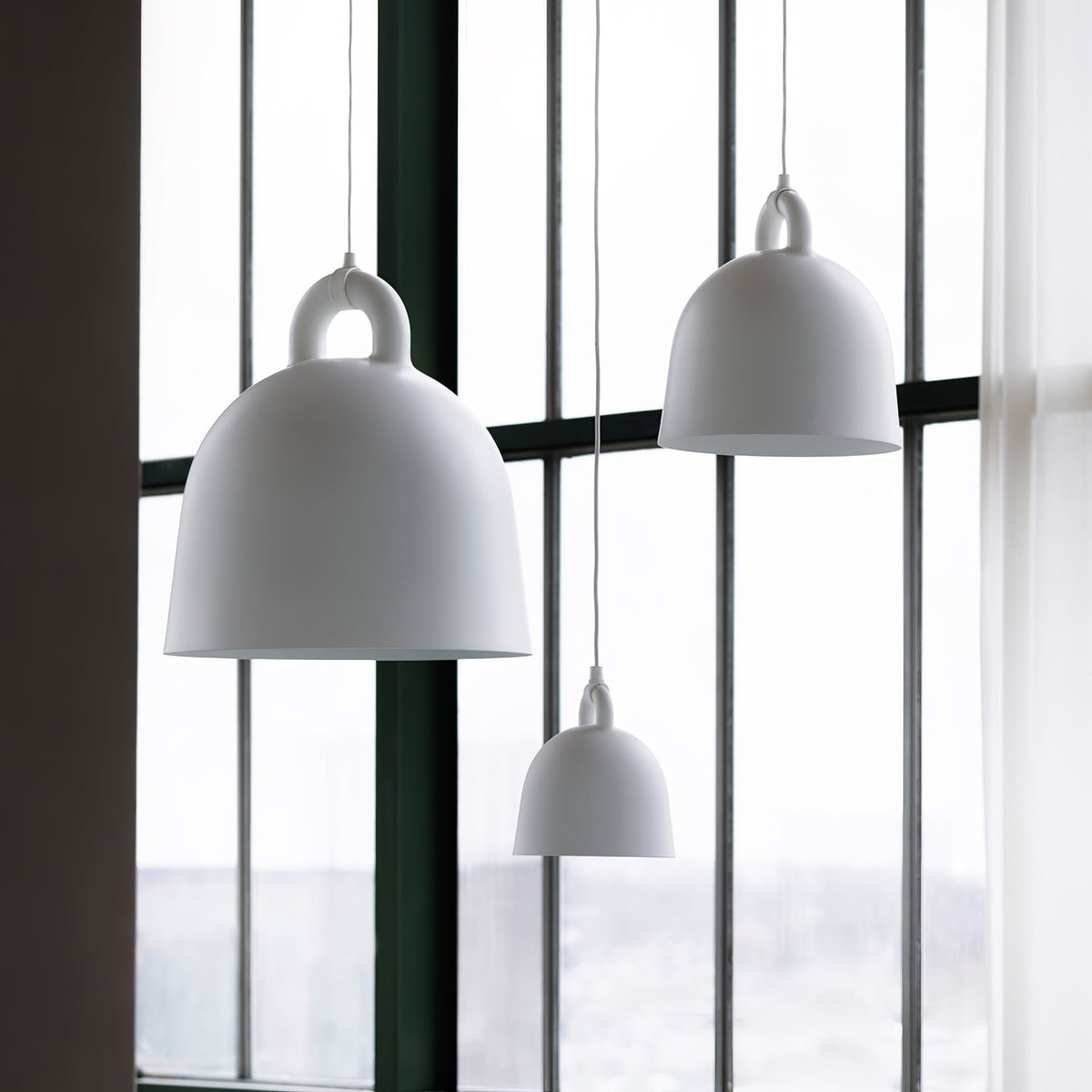 Large and small white Clérachic bell pendants hang by tall windows, with a sheer curtain and a green frame beside them.