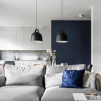 Two black Clérachic bell pendants hang over a grey sofa in a kitchen-living room with a navy panelled wall and blue cushions.