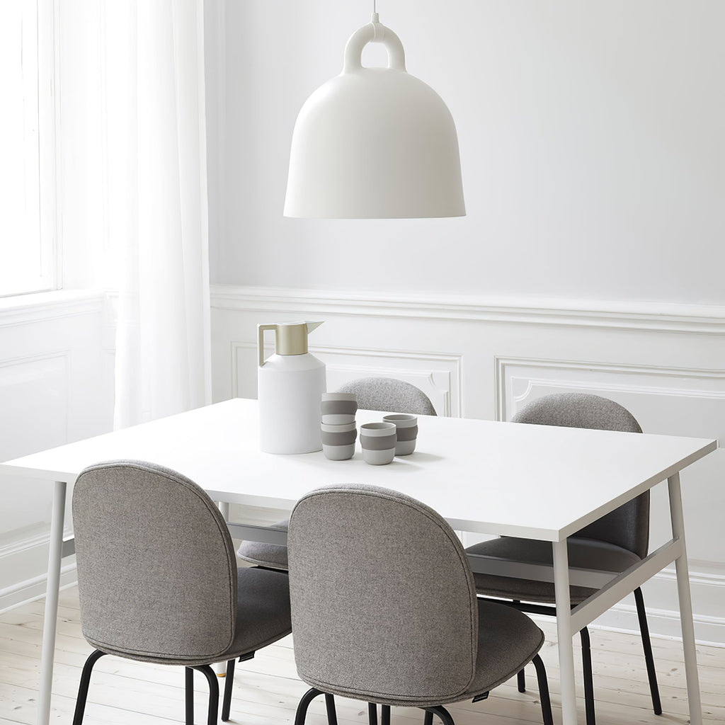 A white Clérachic bell pendant hangs above a white dining table with grey upholstered chairs and a cream jug, in a panelled room.