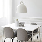 A white Clérachic bell pendant hangs above a white dining table with grey upholstered chairs and a cream jug, in a panelled room.