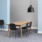 A black Clérachic bell pendant hangs over a long pale oak table with black chairs, in a room of grey carpet and soft blue walls.