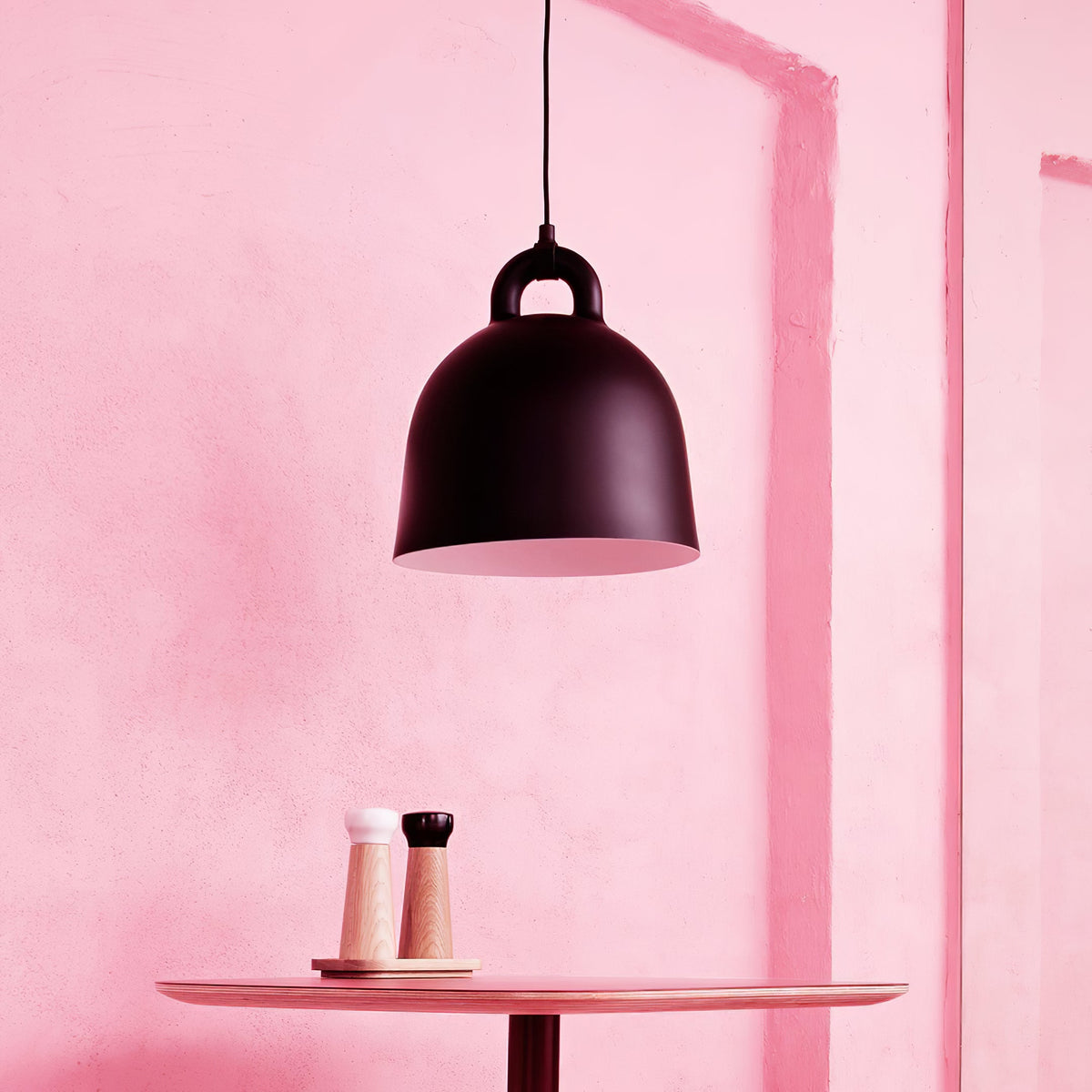A black Clérachic bell pendant hangs over a round pink table set with a pepper mill and a salt mill, against a pink wall.