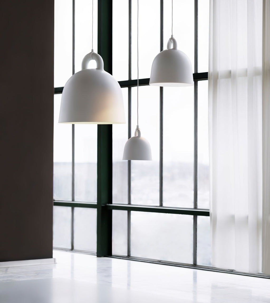 White and grey Clérachic bell pendants hang in front of a tall black-framed window, with a sheer curtain to the right.