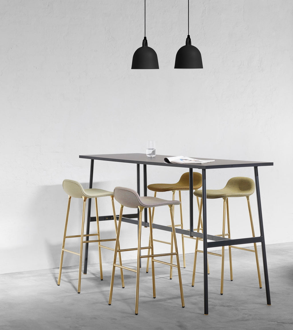Two small black Clérachic bell pendants hang above a tall black table with brass-legged bar stools, against a pale concrete wall.