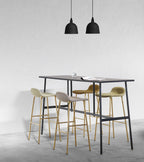 Two small black Clérachic bell pendants hang above a tall black table with brass-legged bar stools, against a pale concrete wall.