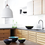 A white Clérachic bell pendant hangs over a kitchen with oak cabinets, a dark worktop, a black tap and colourful bowls.