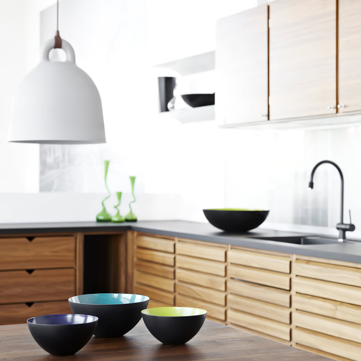 A white Clérachic bell pendant hangs over a kitchen with oak cabinets, a dark worktop, a black tap and colourful bowls.