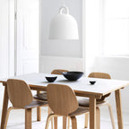 A white Clérachic bell pendant hangs above a white table with wooden chairs, with dark bowls set out on the tabletop.