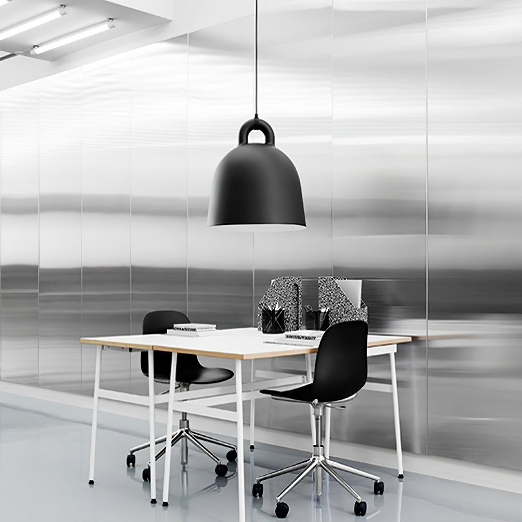 A black Clérachic bell pendant hangs over a white desk with black chairs, in a bright office with brushed metal walls.