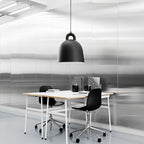 A black Clérachic bell pendant hangs over a white desk with black chairs, in a bright office with brushed metal walls.