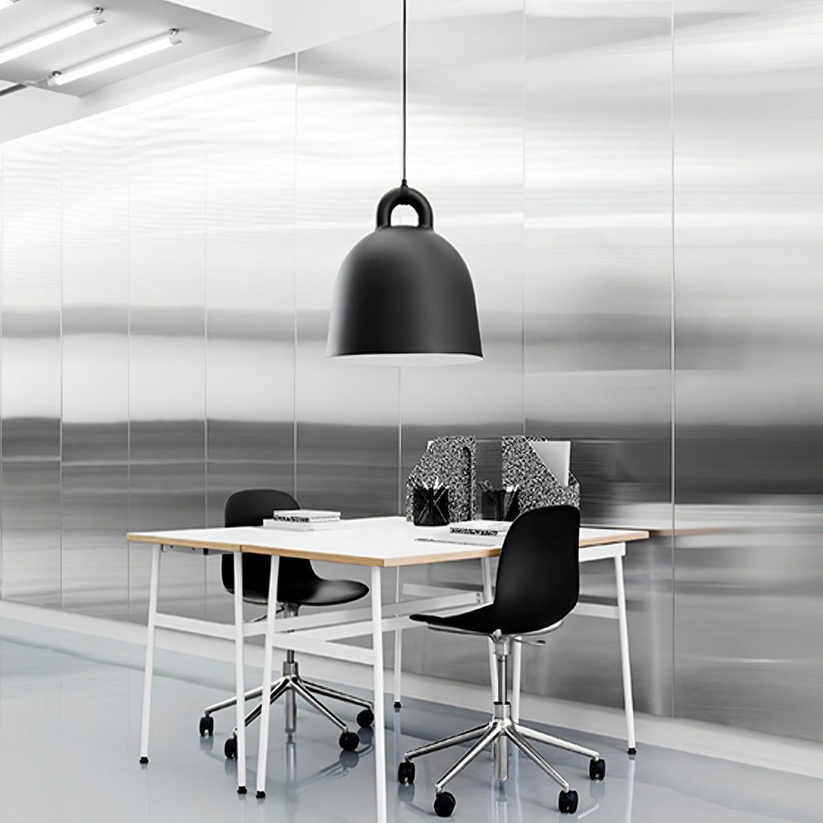 A black Clérachic bell pendant hangs over a white desk with black chairs, in a bright office with brushed metal walls.
