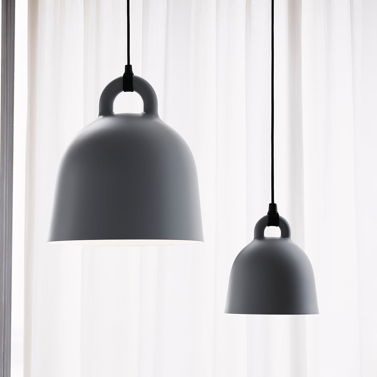 Two grey Clérachic bell pendants of different sizes hang by a sheer white curtain, lit from behind with a soft glow.
