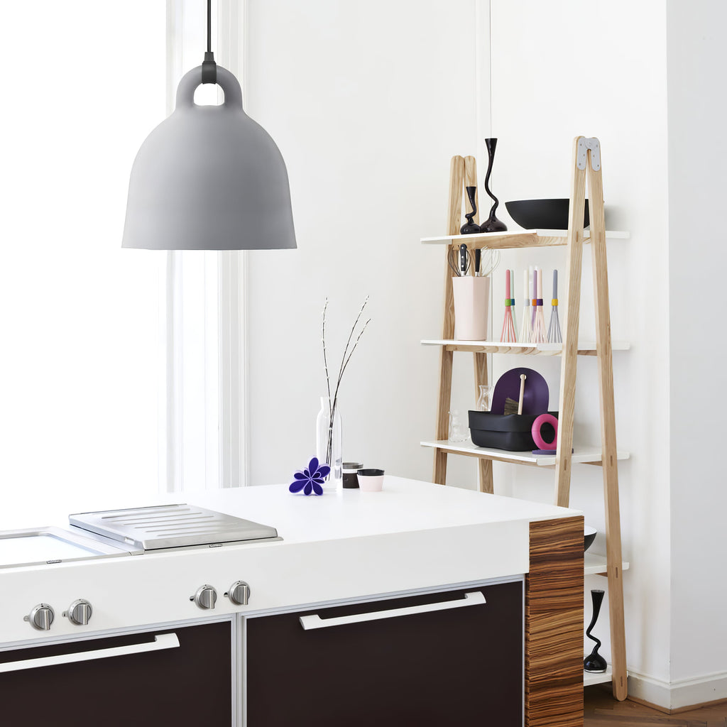 A light grey Clérachic bell pendant hangs by a kitchen island and a ladder-style wooden shelf holding vases and bowls.