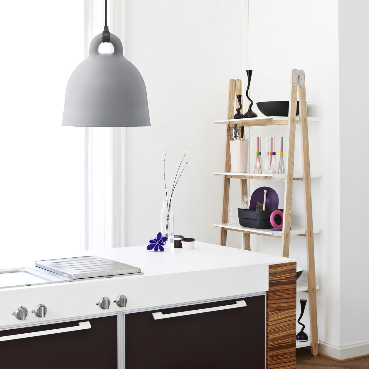 A light grey Clérachic bell pendant hangs by a kitchen island and a ladder-style wooden shelf holding vases and bowls.