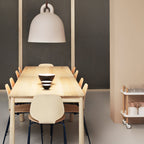 A large sand-coloured Clérachic bell pendant hangs over a long pale wood table, with wooden chairs and a dark mesh wall behind.