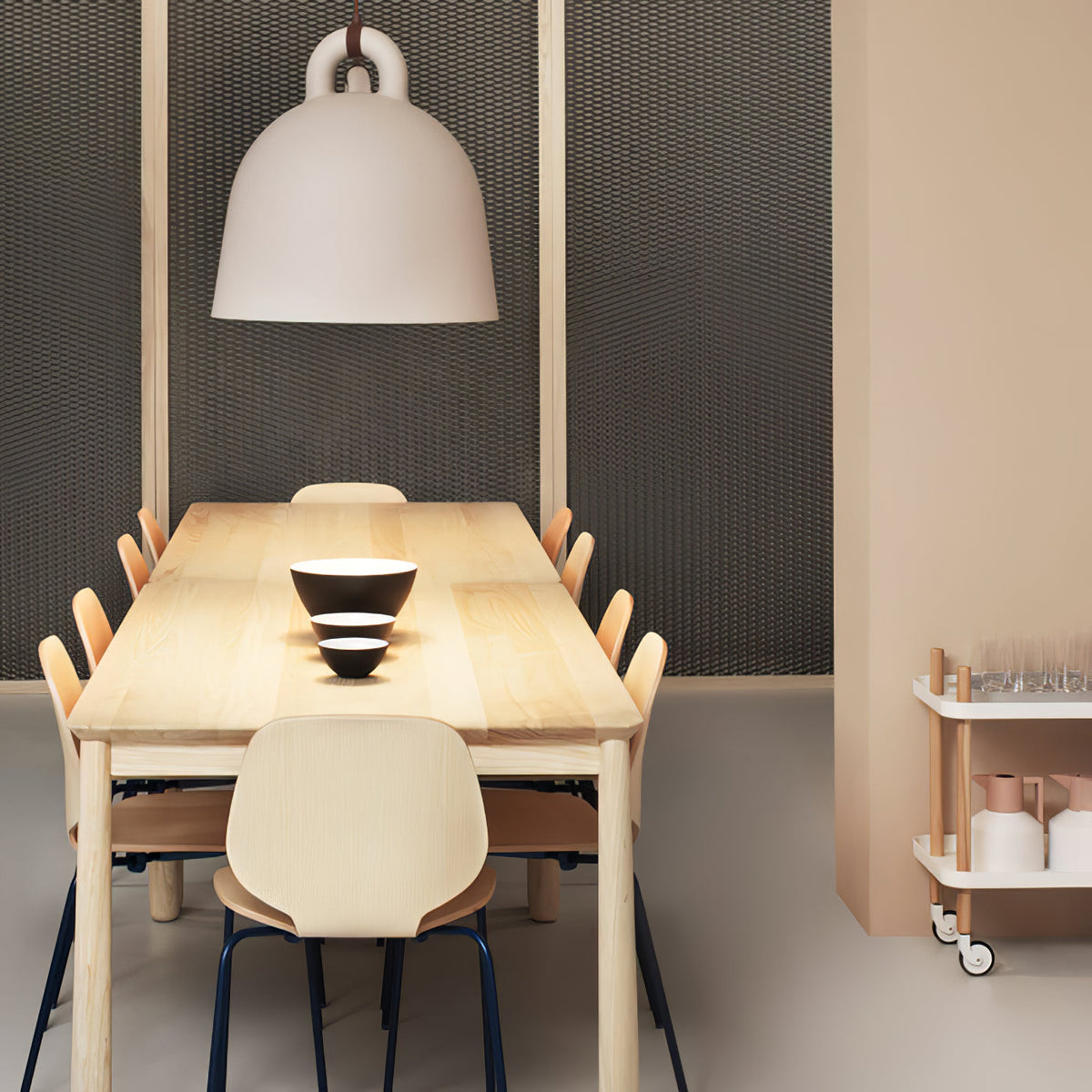 A large sand-coloured Clérachic bell pendant hangs over a long pale wood table, with wooden chairs and a dark mesh wall behind.