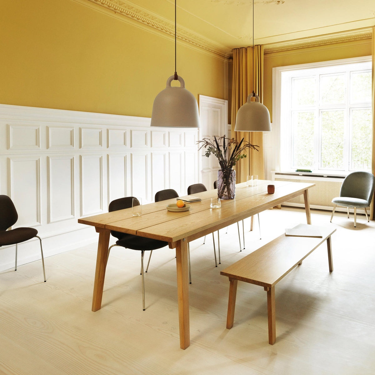 Two Clérachic bell pendants, one grey-taupe, hang over a long oak table in a high-ceilinged room with yellow walls and white panelling.