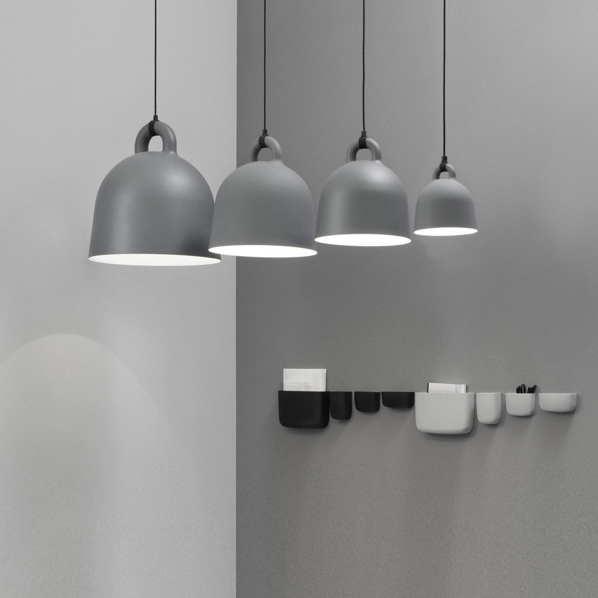 Four grey Clérachic bell pendants in graduated sizes hang on a grey wall, above a row of black and white wall pockets.