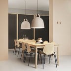Two sand-coloured Clérachic bell pendants hang over a long pale wood table, with a peach wall panel and dark mesh behind.