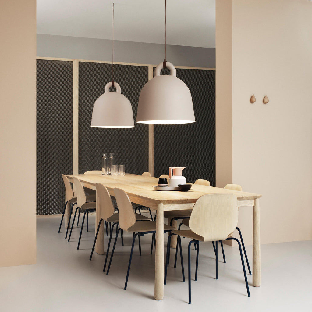Two sand-coloured Clérachic bell pendants hang over a pale wood dining table, set against a dark mesh wall and peach panels.