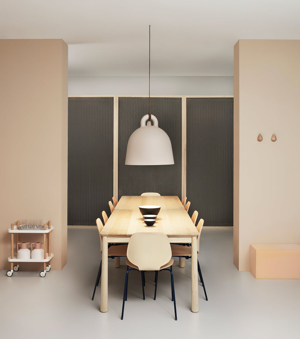 A sand-coloured Clérachic bell pendant hangs at the end of a long wood table, between peach walls, with a trolley to the left.