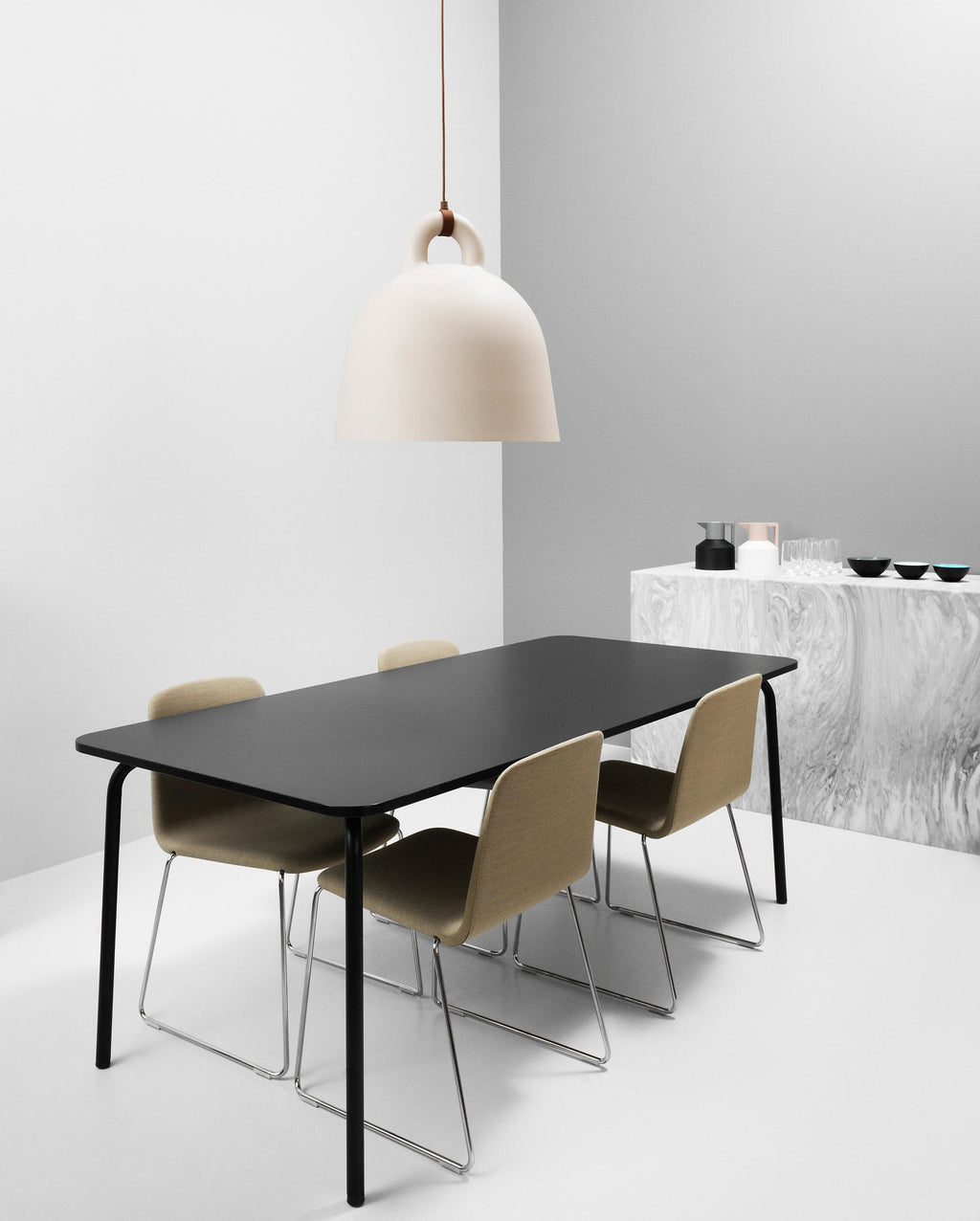 A cream Clérachic bell pendant with a brown cord hangs by a white and grey wall, with a black table and olive chairs below.