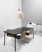 A cream Clérachic bell pendant with a brown cord hangs by a white and grey wall, with a black table and olive chairs below.