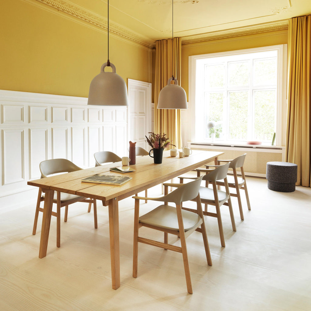 Two sand-coloured Clérachic bell pendants hang over a wooden table in a yellow room with white panelling and a window.