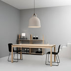 A sand-coloured Clérachic bell pendant hangs in a grey room with a pale wood table, black chairs and a shelving unit.