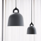 Two grey Clérachic bell pendants, one large and one small, hang before a sheer curtain, shown close up from below.