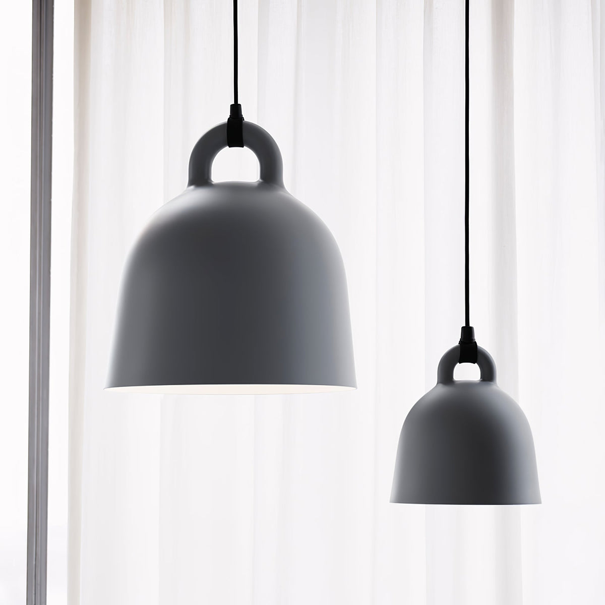 Two grey Clérachic bell pendants, one large and one small, hang before a sheer curtain, shown close up from below.