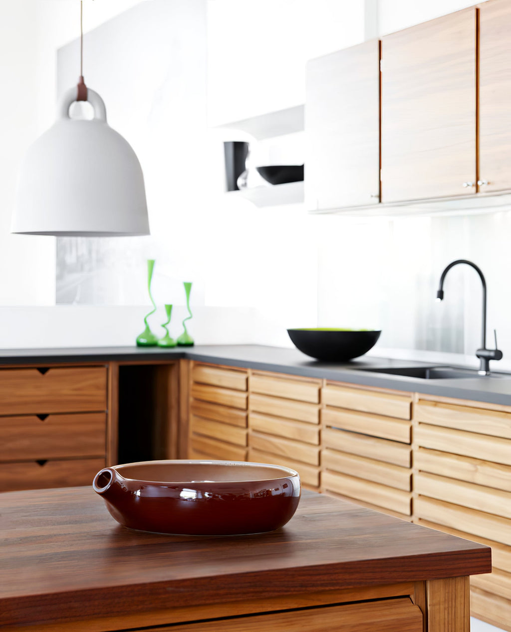 A white Clérachic bell pendant hangs over a kitchen with oak cabinets, a dark worktop and a red-brown bowl on the counter.