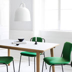 A white Clérachic bell pendant hangs above a white table with green chairs, near a bright window with a candle on the table.