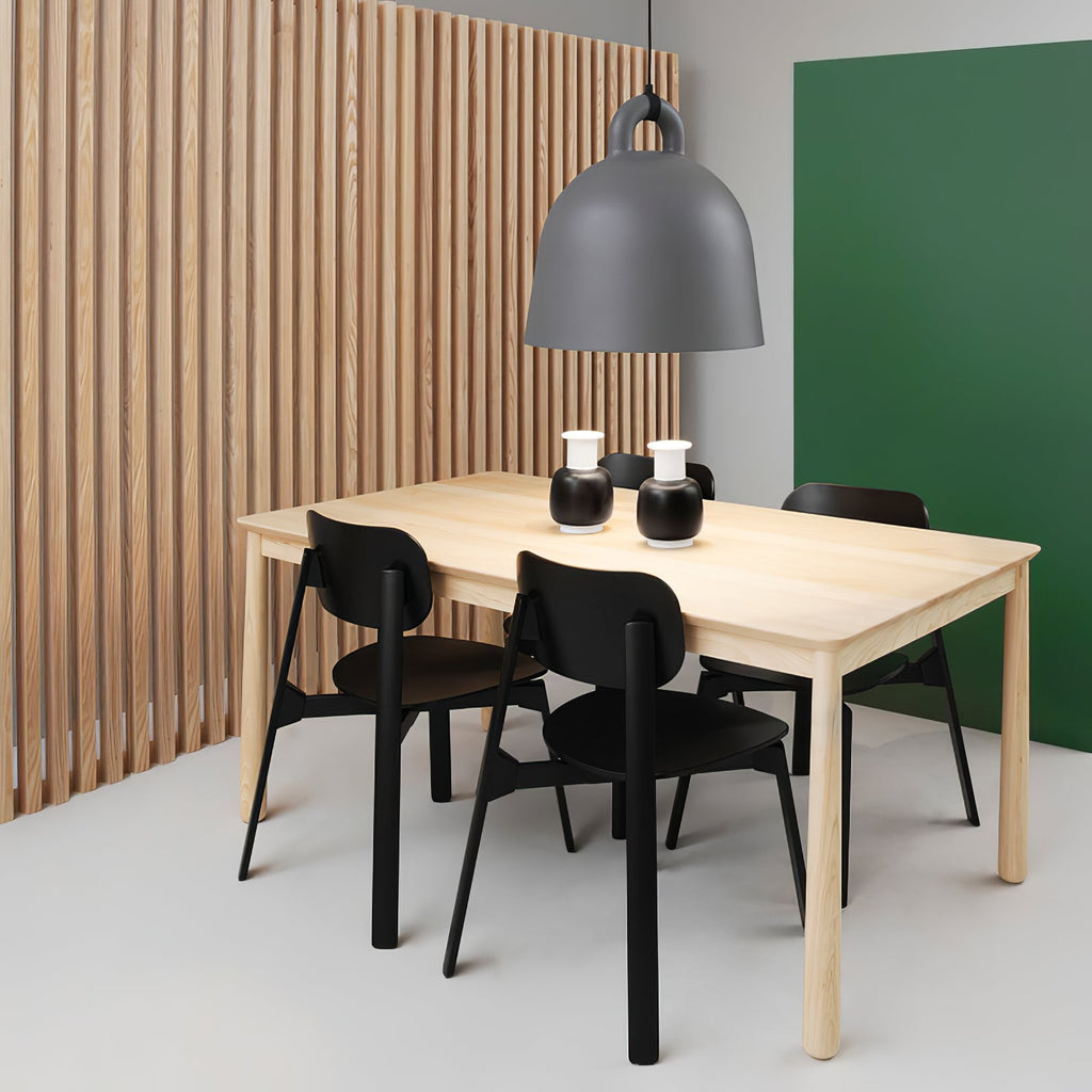 A grey Clérachic bell pendant hangs over a pale wood table with black chairs, in front of wood slats and a green wall.