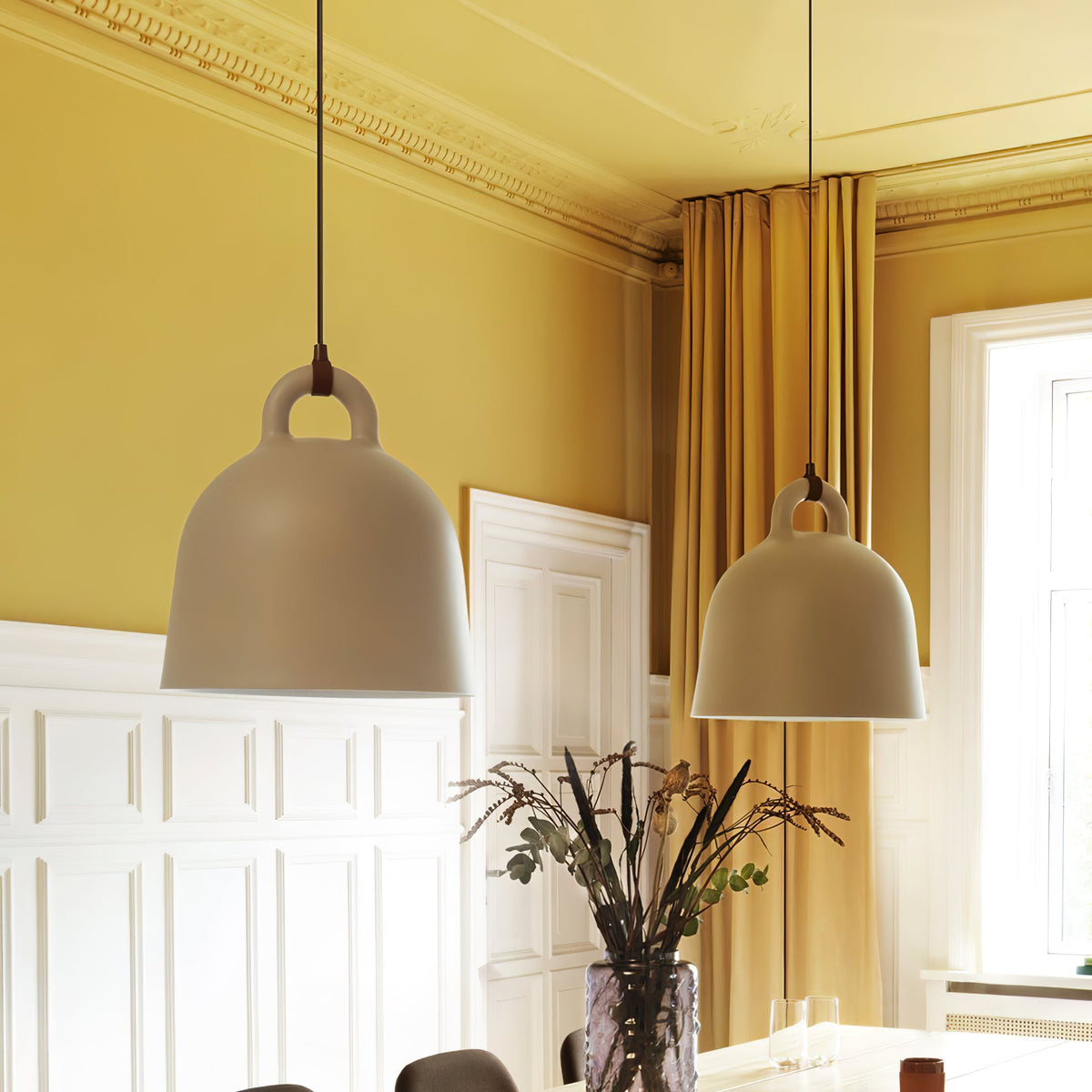 Two sand-coloured Clérachic bell pendants hang in a room with yellow walls, white panelling and a dried-flower vase below.