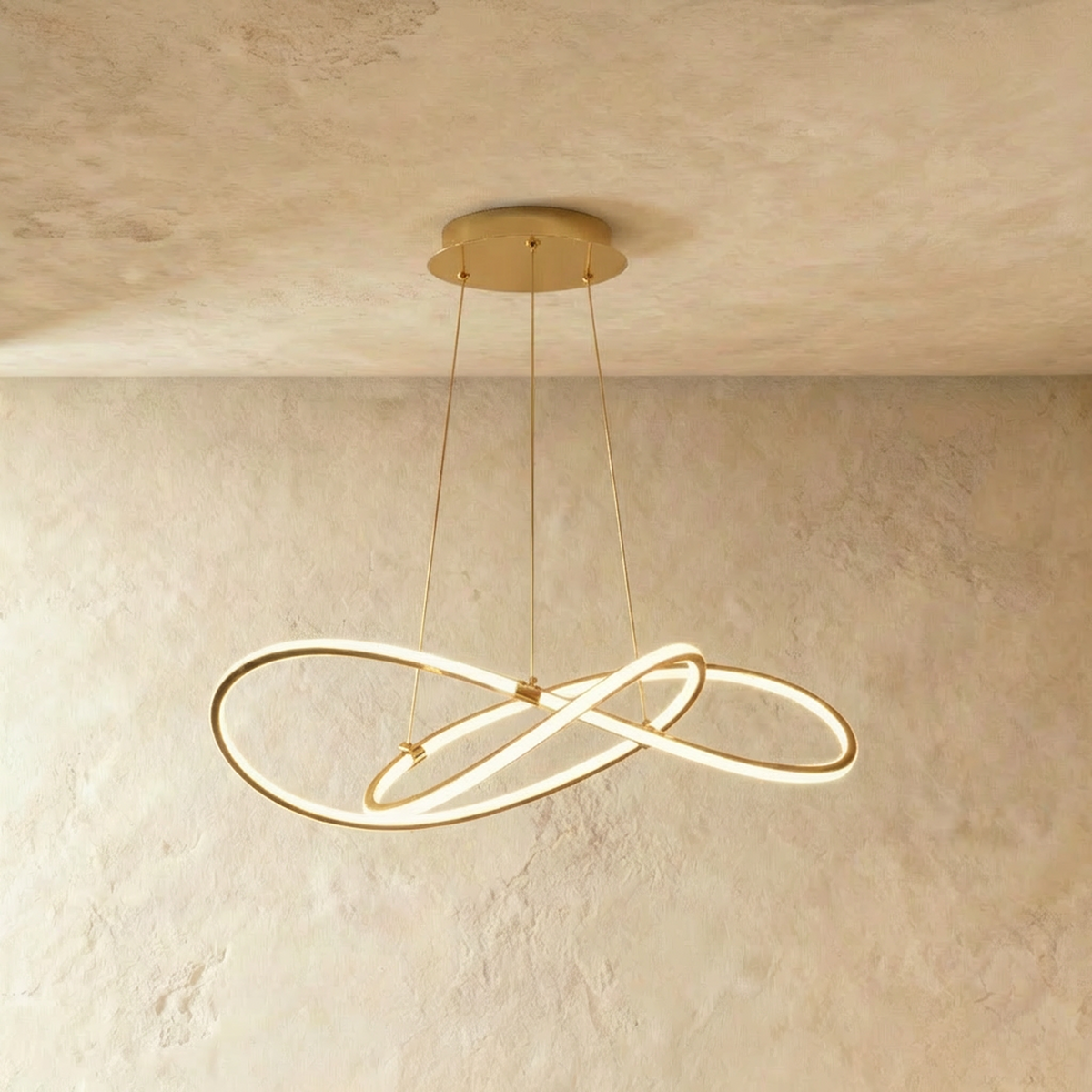 Clérachic brass-finish pendant of interlaced LED loops on three slim brass cables, lit against a textured plaster wall.