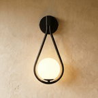 Clérachic black iron arm wall sconce with a teardrop frame cradling a lit opal glass globe on a plaster wall.